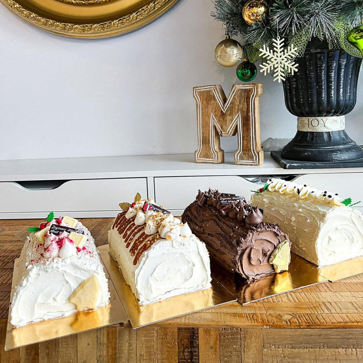 Three decorated desserts on a wooden board with a Christmas tree and decorative mirror in the background.