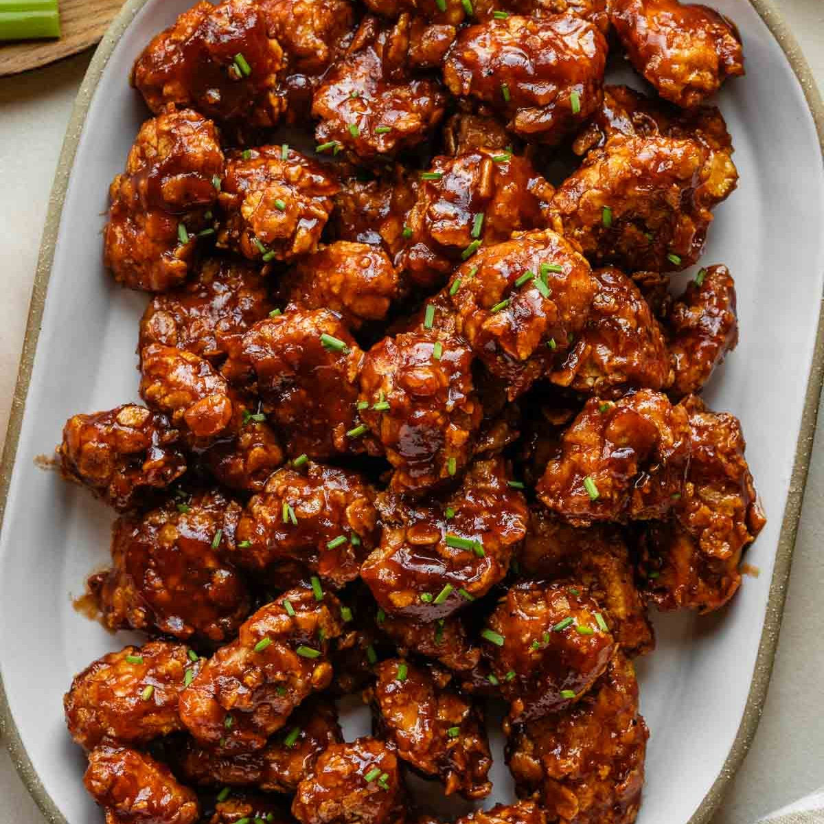 Succulent glazed chicken tenders coated in a glossy reddish-brown sauce and topped with chopped chives, served on a large oval white platter.