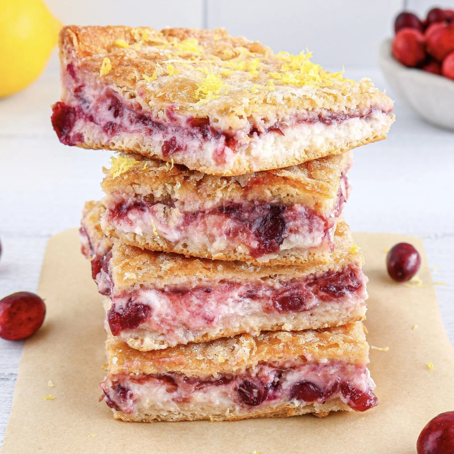 Stack of cranberry lemon bars on a white surface with a lemon and cranberries in the background.