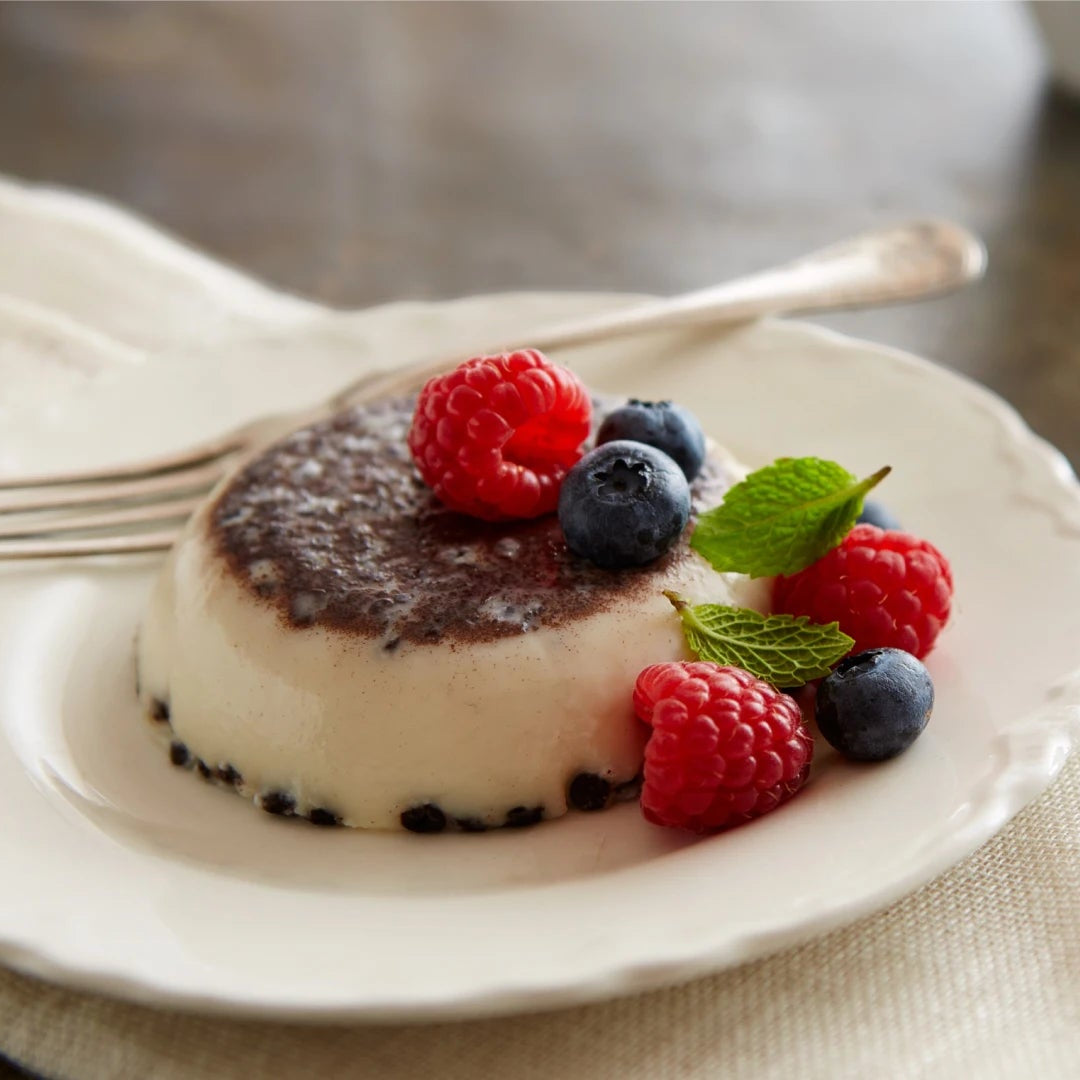 A small dessert panna cotta on a white plate, topped with fresh raspberries, blueberries, mint leaves, and a dusting of powdered sugar, set on a beige cloth with a fork and blurred chocolate kisses in the background.