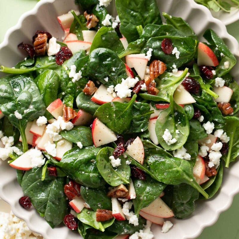 Spinach salad with apples, cranberries, and feta cheese in a white bowl on a green background