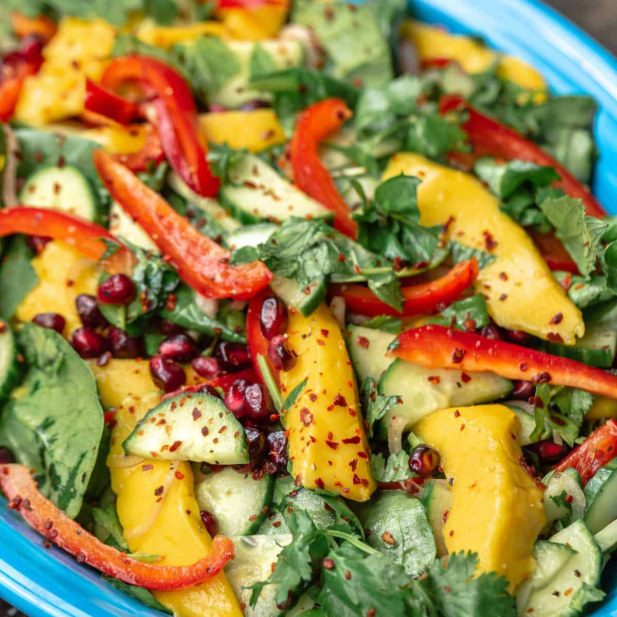 A close-up of a colorful fruit and vegetable salad in a bright blue bowl, featuring yellow mango pieces, red bell pepper strips, cucumber slices, and fresh green herbs on a dark rustic surface.
