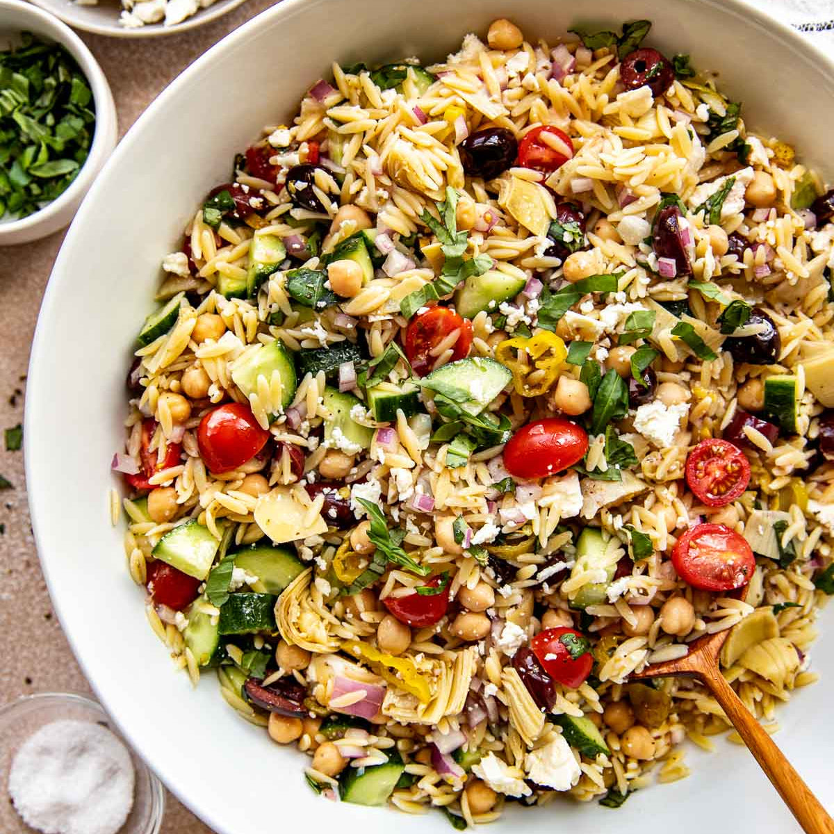 A large white bowl filled with a colorful Mediterranean orzo salad, surrounded by ingredients including chickpeas, cherry tomatoes, cucumber, feta cheese, parsley, salt, pepper, and a lemon wedge on a neutral tabletop.