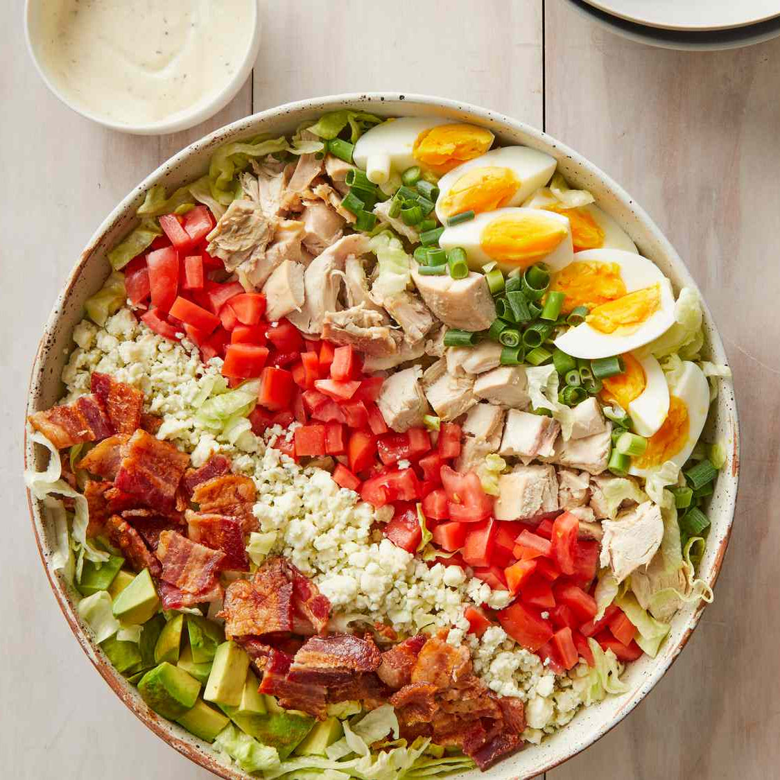 Large salad with chicken, bacon, eggs, and vegetables in a white bowl on a wooden surface.