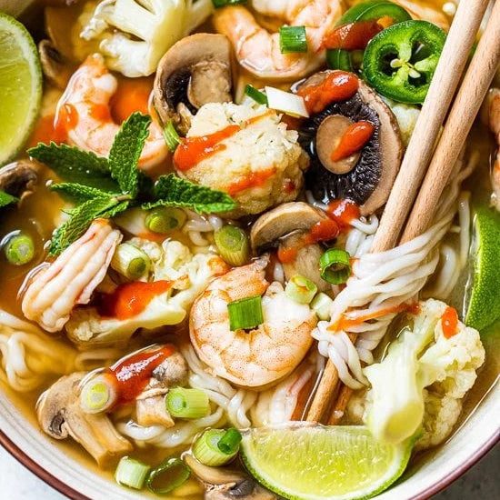 bowl of noodle soup with shrimp, vegetables, and lime wedges