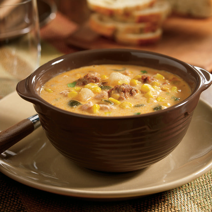 Brown bowl of creamy corn chowder with sausage on a beige plate.