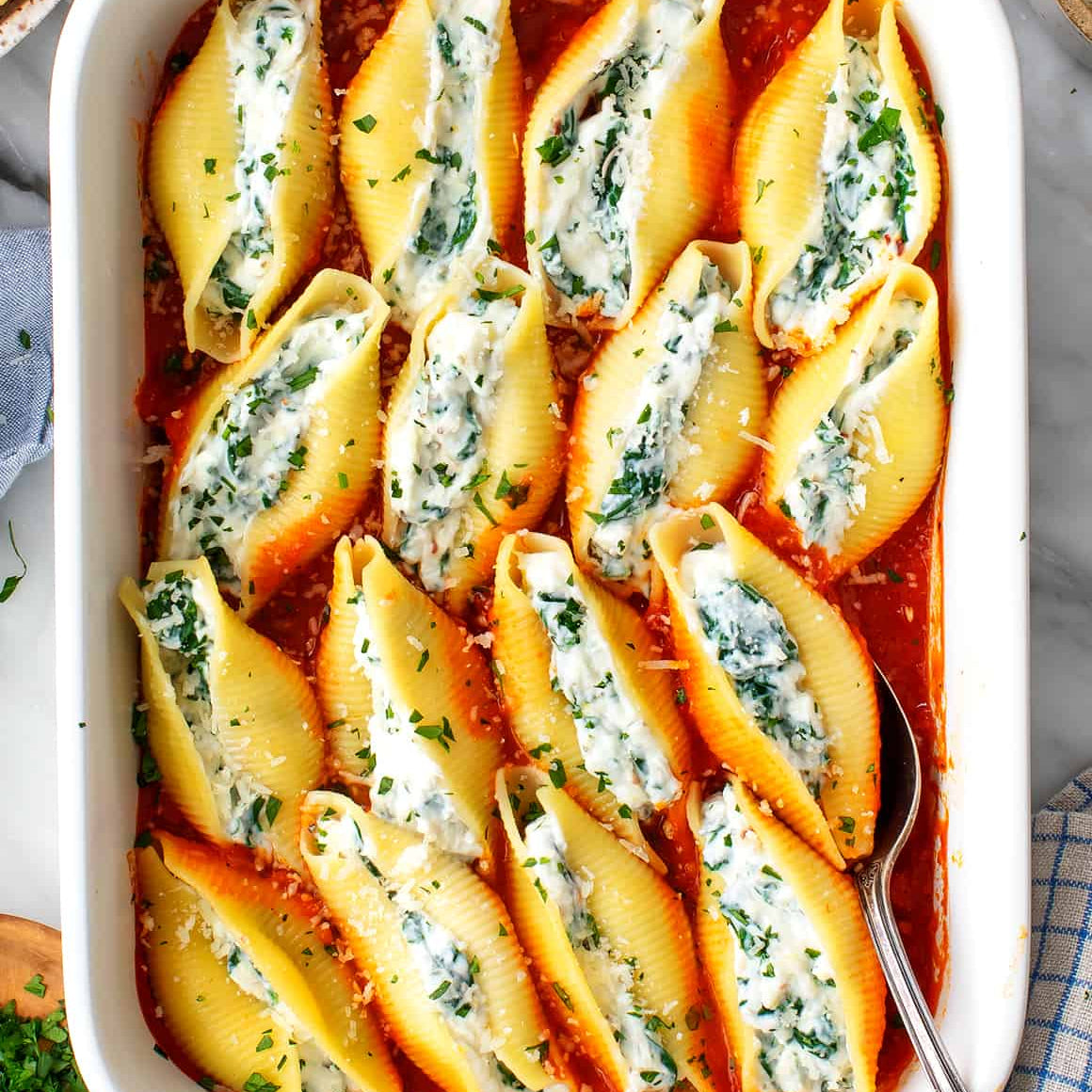Stuffed pasta shells in a white baking dish with a spoon on a checkered cloth.