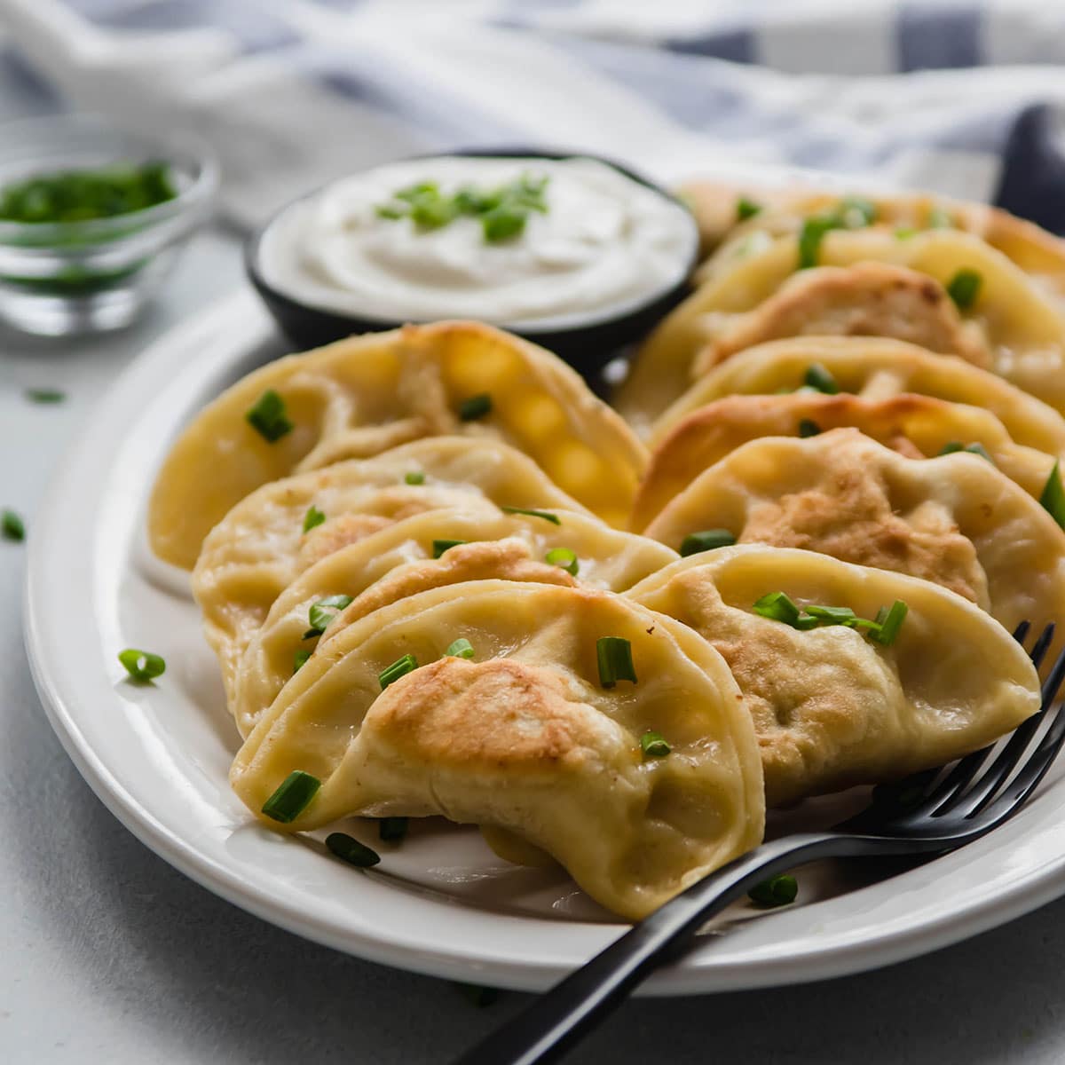 Plated dumplings with a side of sour cream and chives on a checkered tablecloth.