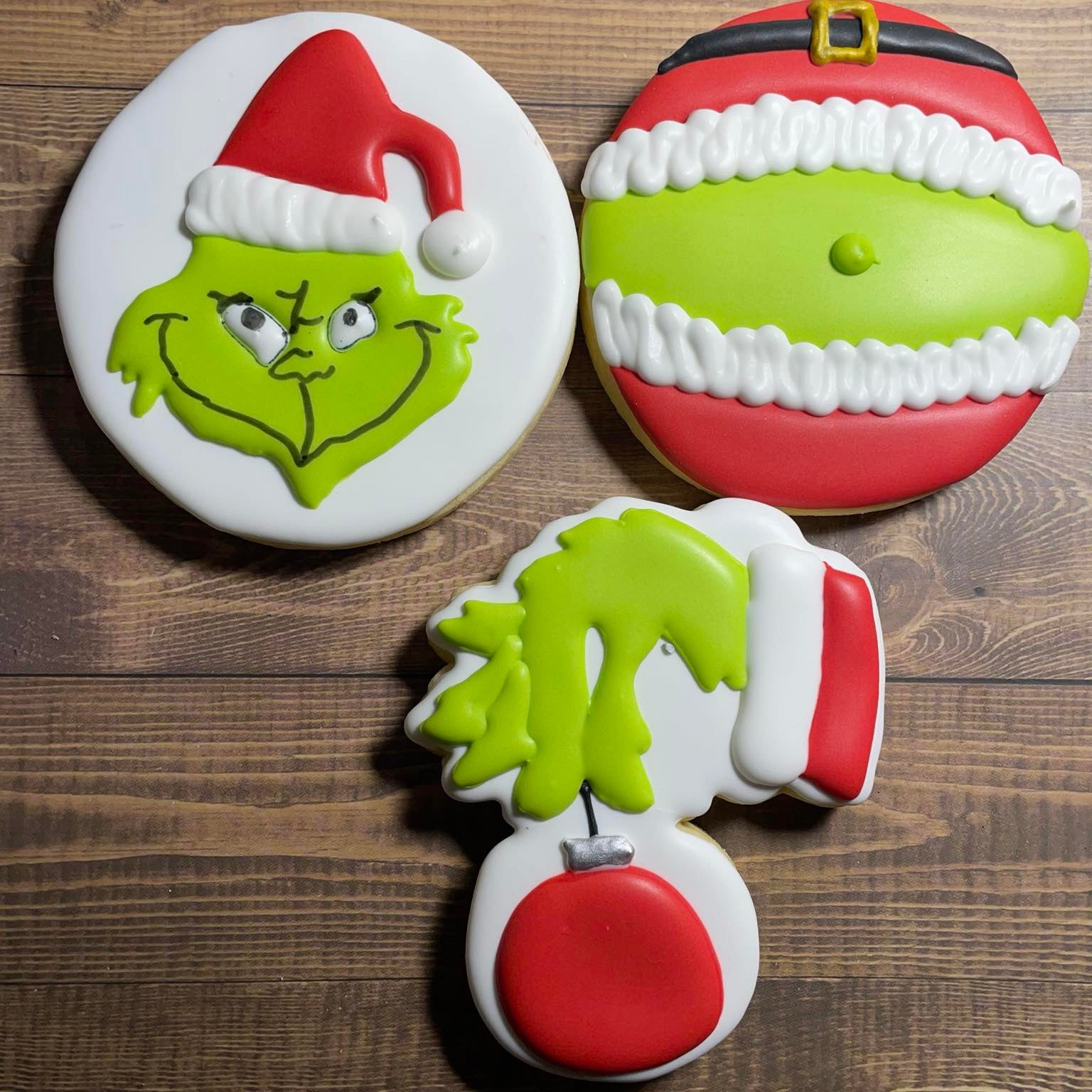 Three cookies shaped like the Grinch with a Santa hat on a wooden surface
