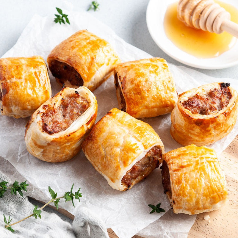 Sausage rolls on a white surface with a small dish of honey and a wooden dipper.