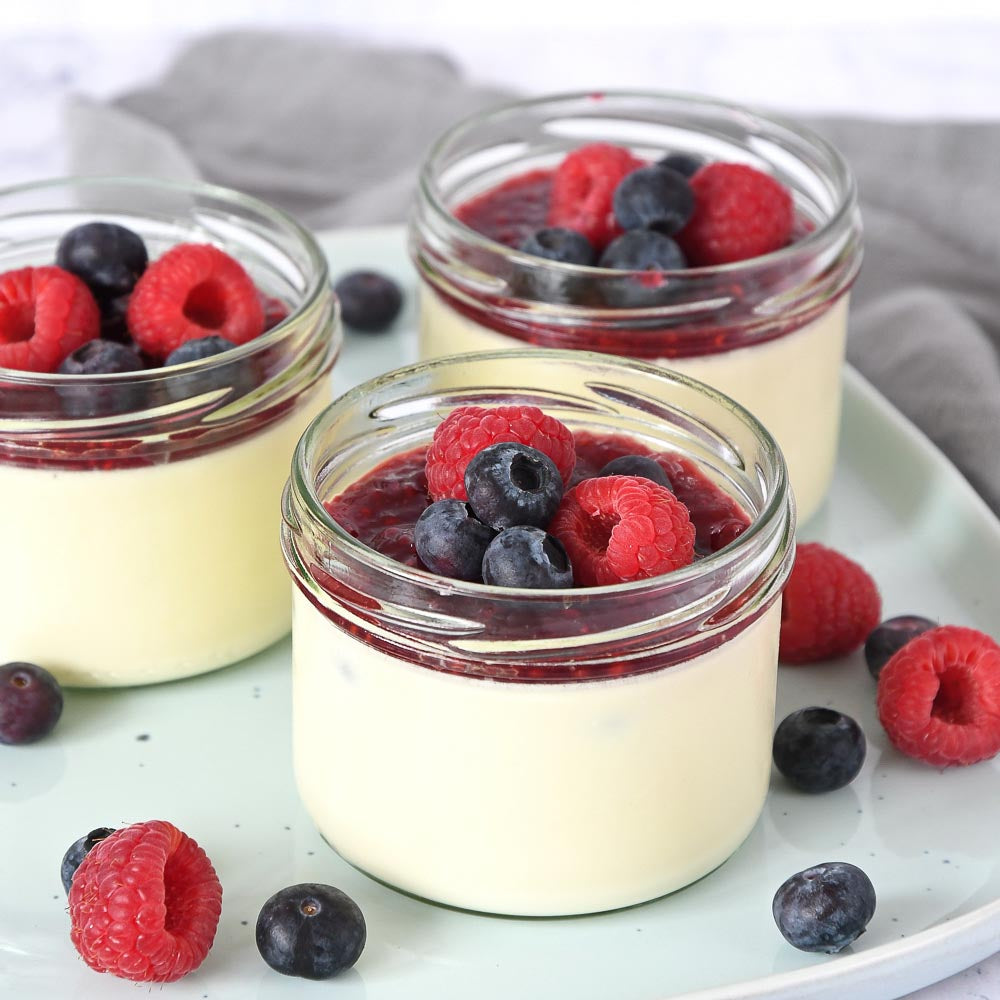 Three small glass jars of creamy custard dessert topped with fresh raspberries and blueberries, arranged on a pale plate with additional berries scattered around.