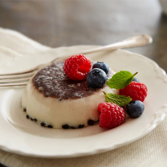 A small dessert panna cotta on a white plate, topped with fresh raspberries, blueberries, mint leaves, and a dusting of powdered sugar, set on a beige cloth with a fork and blurred chocolate kisses in the background.