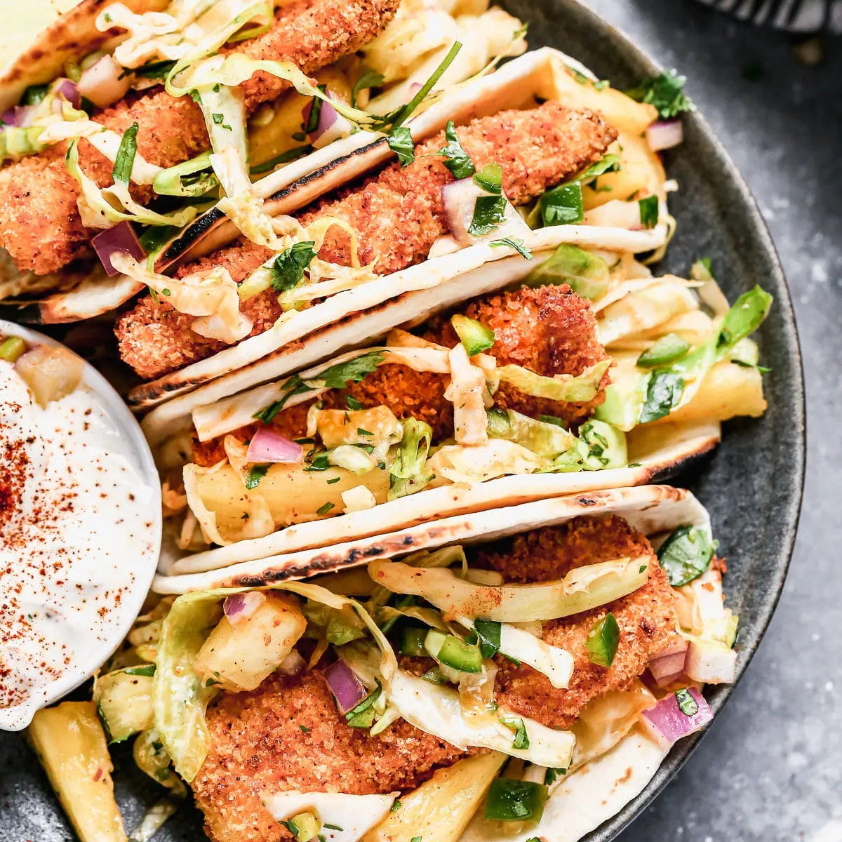 Three fish tacos filled with breaded fish fillets, shredded cabbage, red onion, and cilantro, served on a dark plate with a side of sour cream and lime wedges on a gray surface.