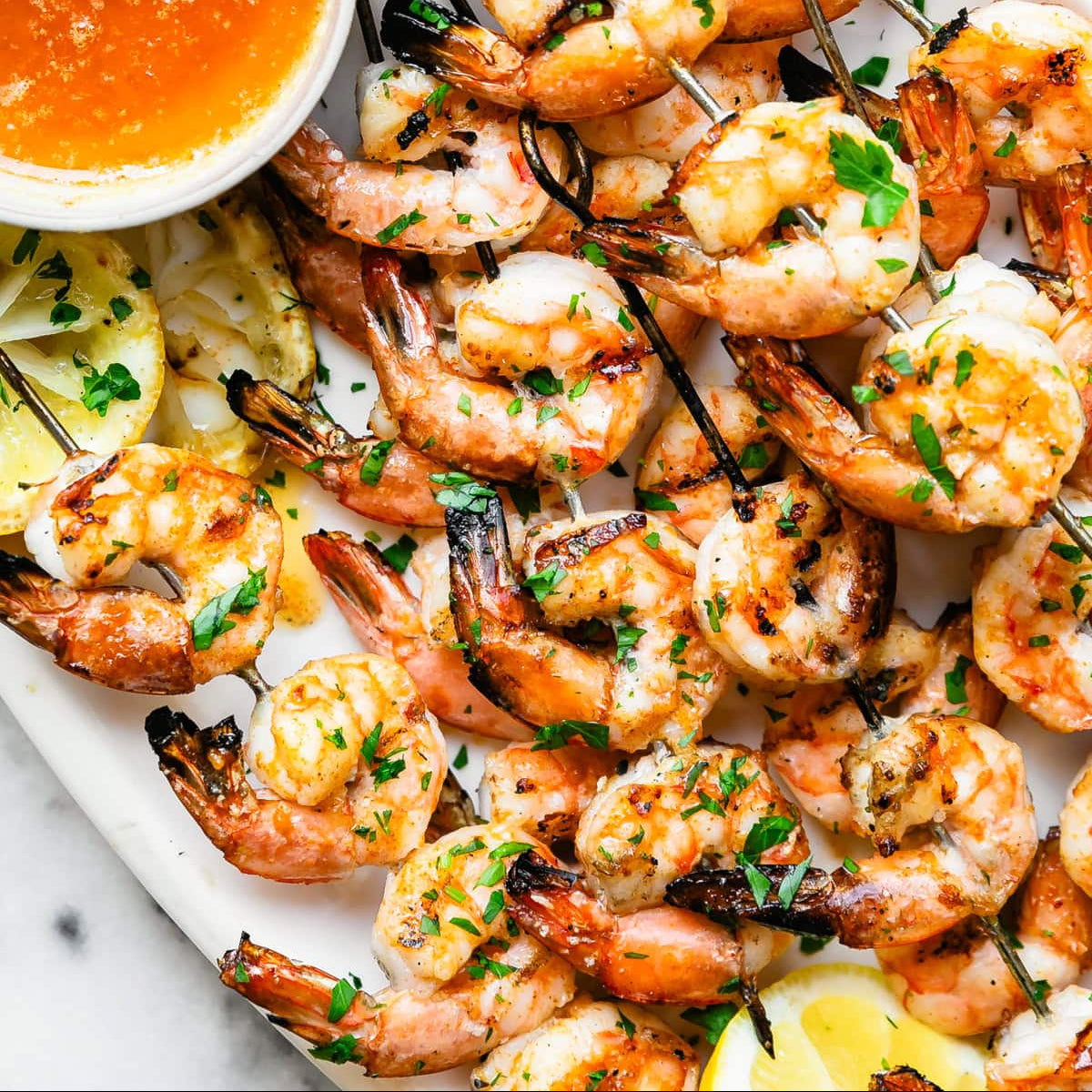 Grilled shrimp skewers garnished with chopped parsley, served on a white platter with a small bowl of orange dipping sauce and lemon wedges.