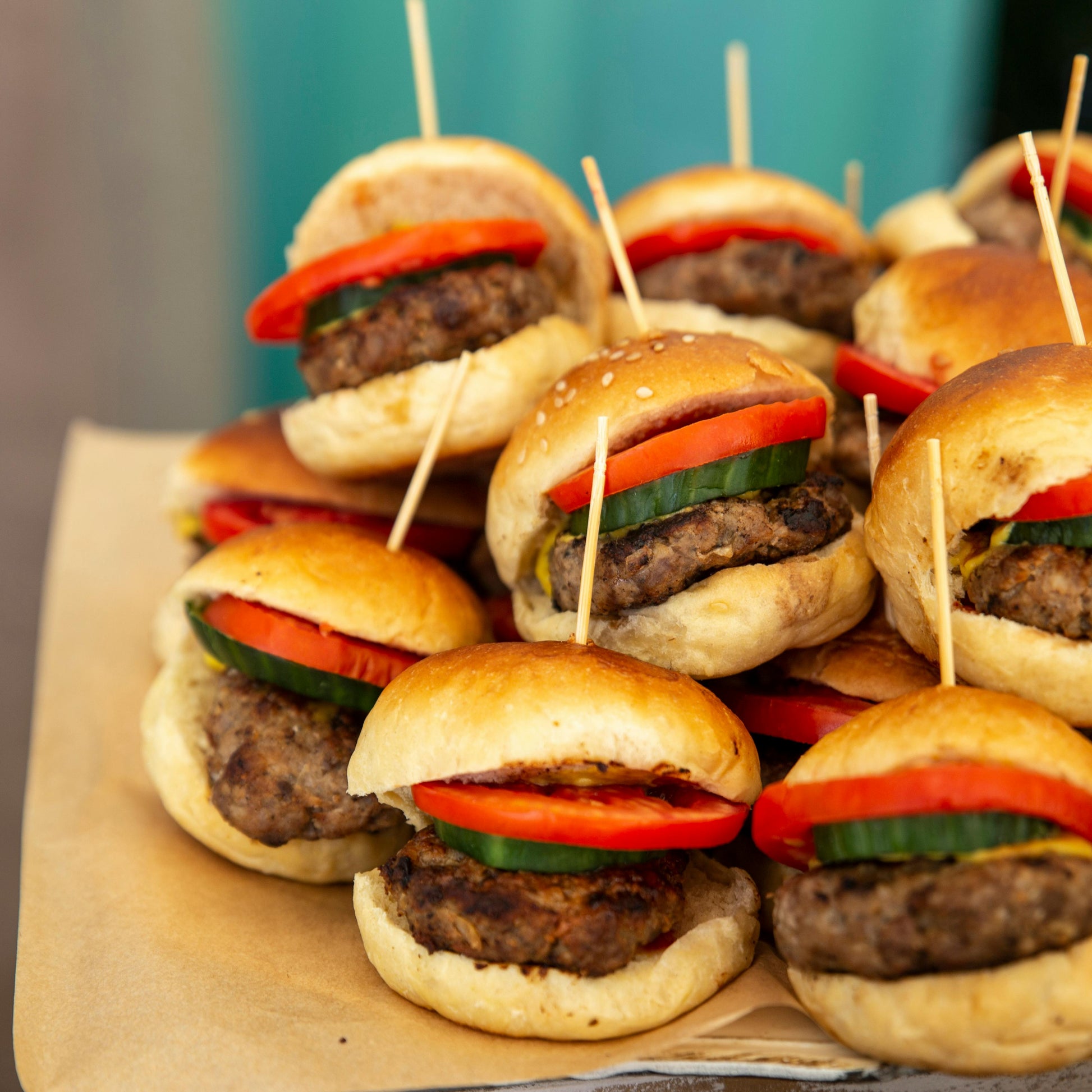 Stack of mini burgers with buns, tomatoes, and cucumbers on a wooden board.
