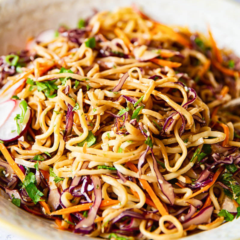 A close-up shot of a fresh Asian noodle salad in a light beige ceramic bowl. The salad features golden-brown noodles mixed with shredded purple cabbage, carrots, radishes, and green cilantro.