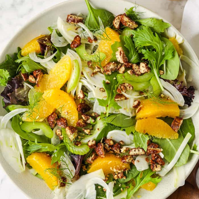 Salad with oranges, nuts, and greens in a white bowl on a marble surface.