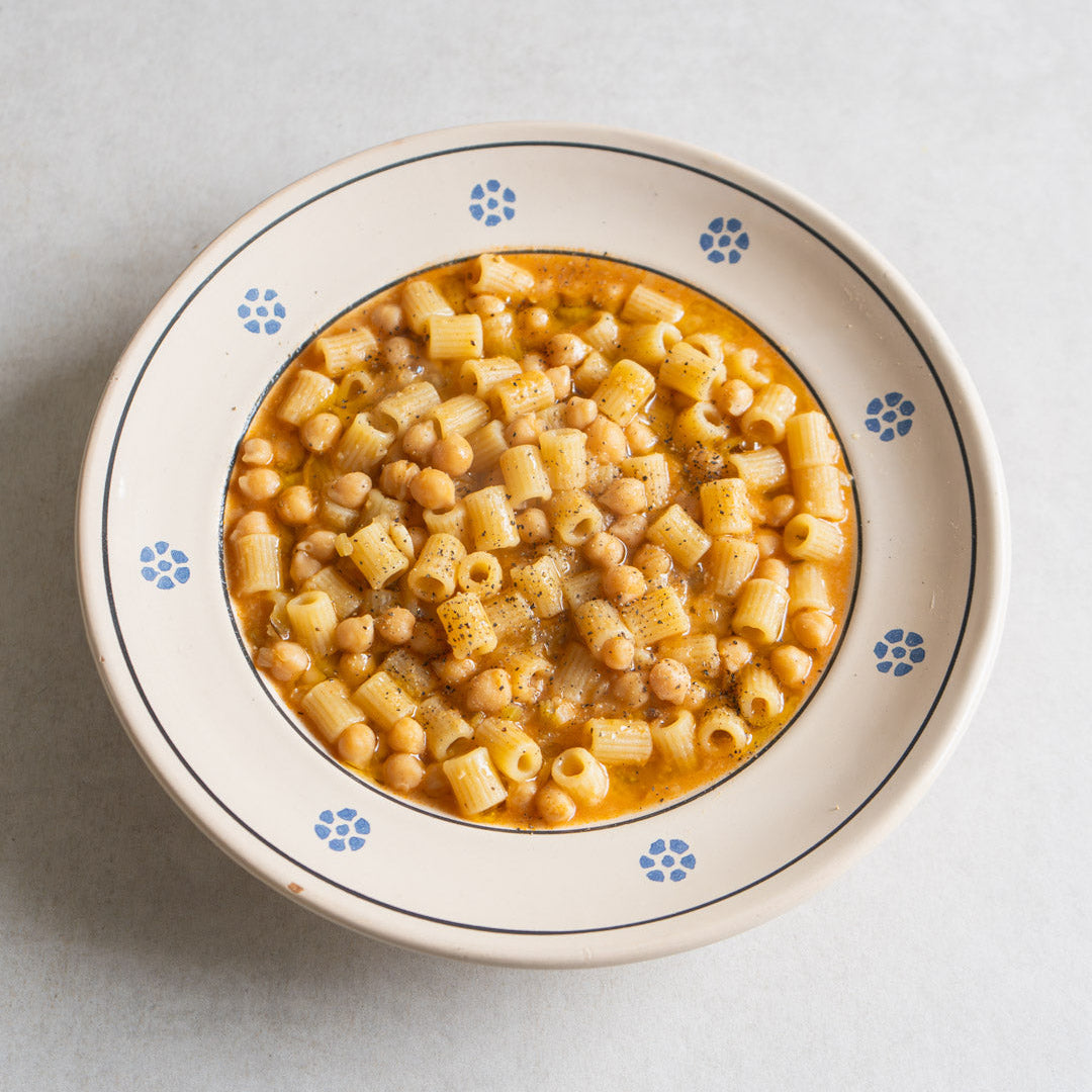 A top-down view of a bowl of macaroni pasta with chickpeas in a light orange sauce, served in a cream-colored ceramic bowl with blue floral accents on a light gray background.