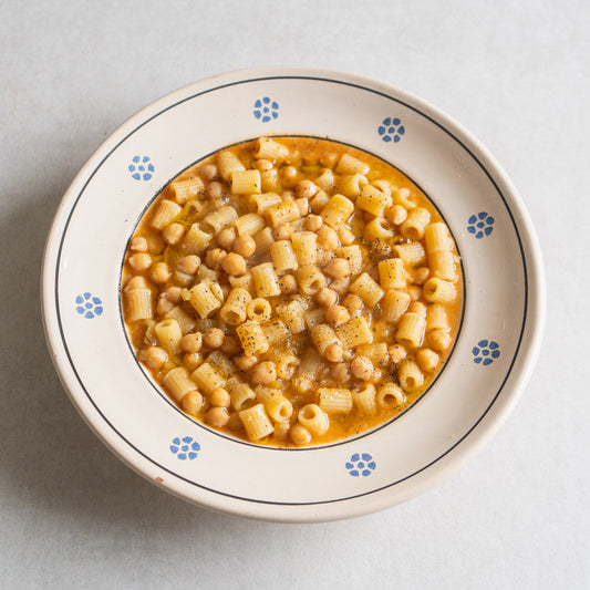 A top-down view of a bowl of macaroni pasta with chickpeas in a light orange sauce, served in a cream-colored ceramic bowl with blue floral accents on a light gray background.