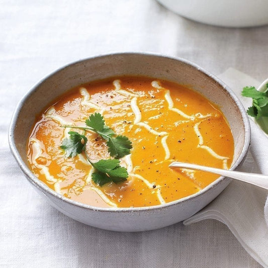 A bowl of orange soup, likely pumpkin or butternut squash, garnished with a swirl of white cream and fresh cilantro, served in a speckled gray ceramic bowl on a white textured tablecloth.