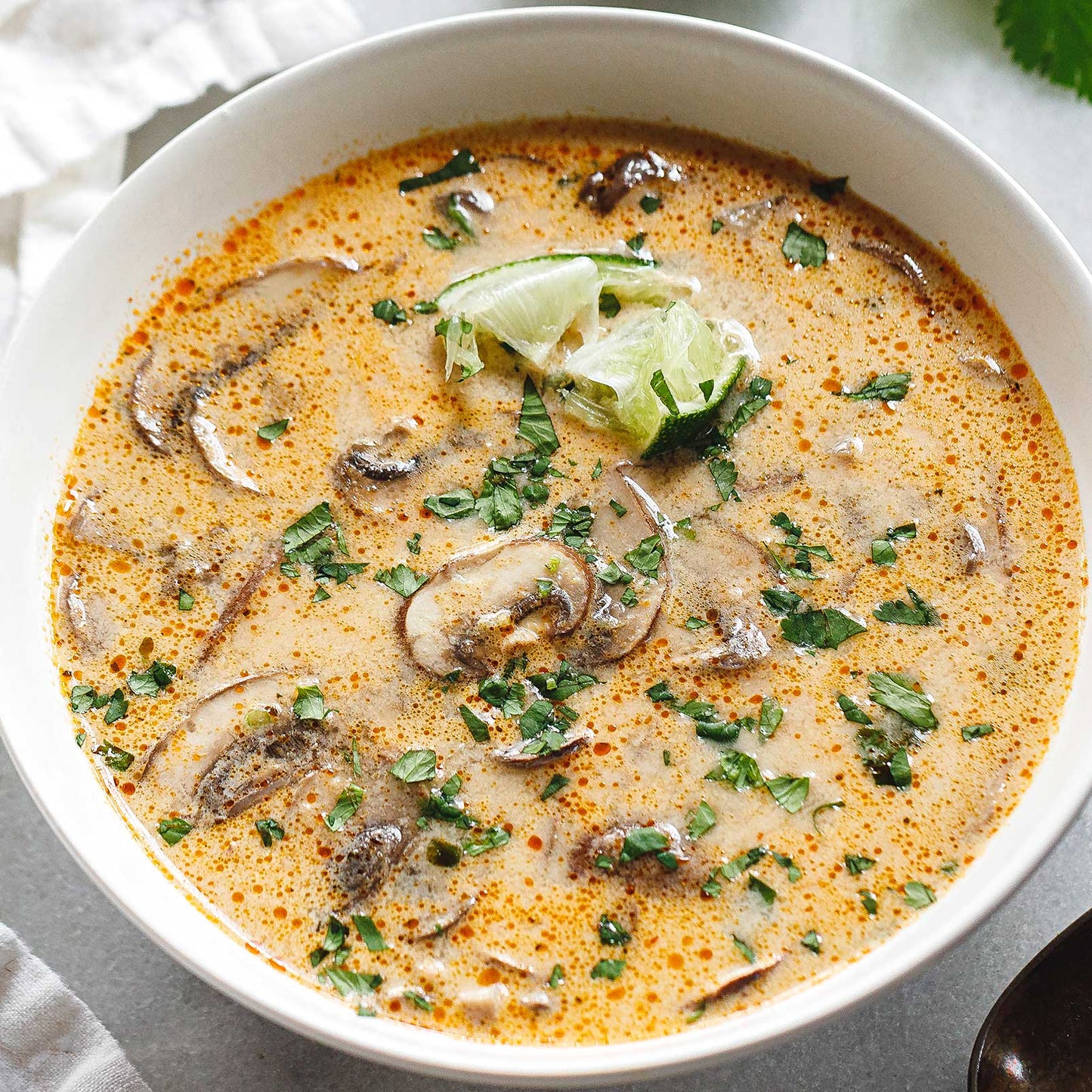 A white ceramic bowl filled with creamy mushroom soup topped with chopped green herbs, sliced mushrooms, and a lime wedge, set on a light gray surface with a spoon and white cloth nearby.