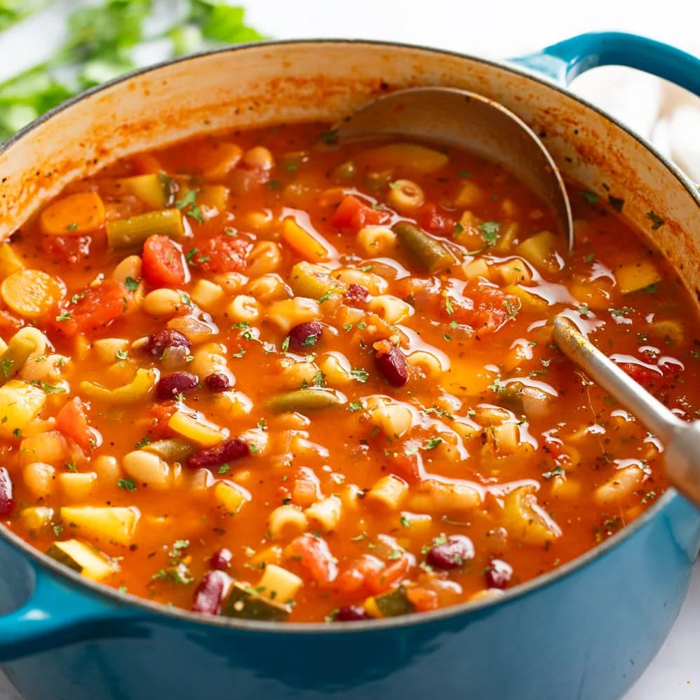 Pasta soup in a blue pot with a spoon on a marble surface