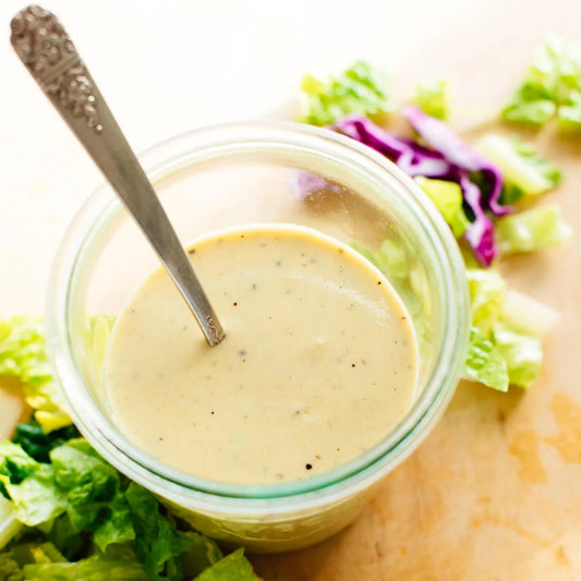 Jar of creamy salad dressing with a spoon, surrounded by lettuce on a wooden surface.