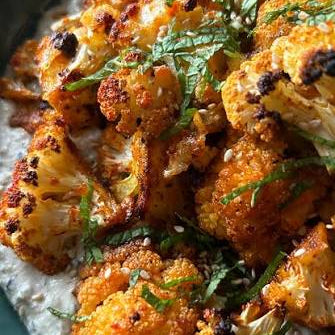 Roasted cauliflower florets with a golden-brown crispy exterior, sprinkled with herbs and sesame seeds, served over a creamy white dip in a dark teal bowl.
